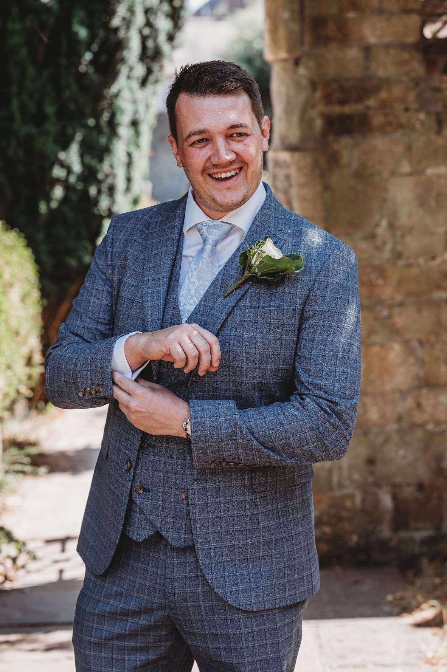 A person wearing a gray windowpane suit with a boutonniere smiles while adjusting their cuff on a sunny day.