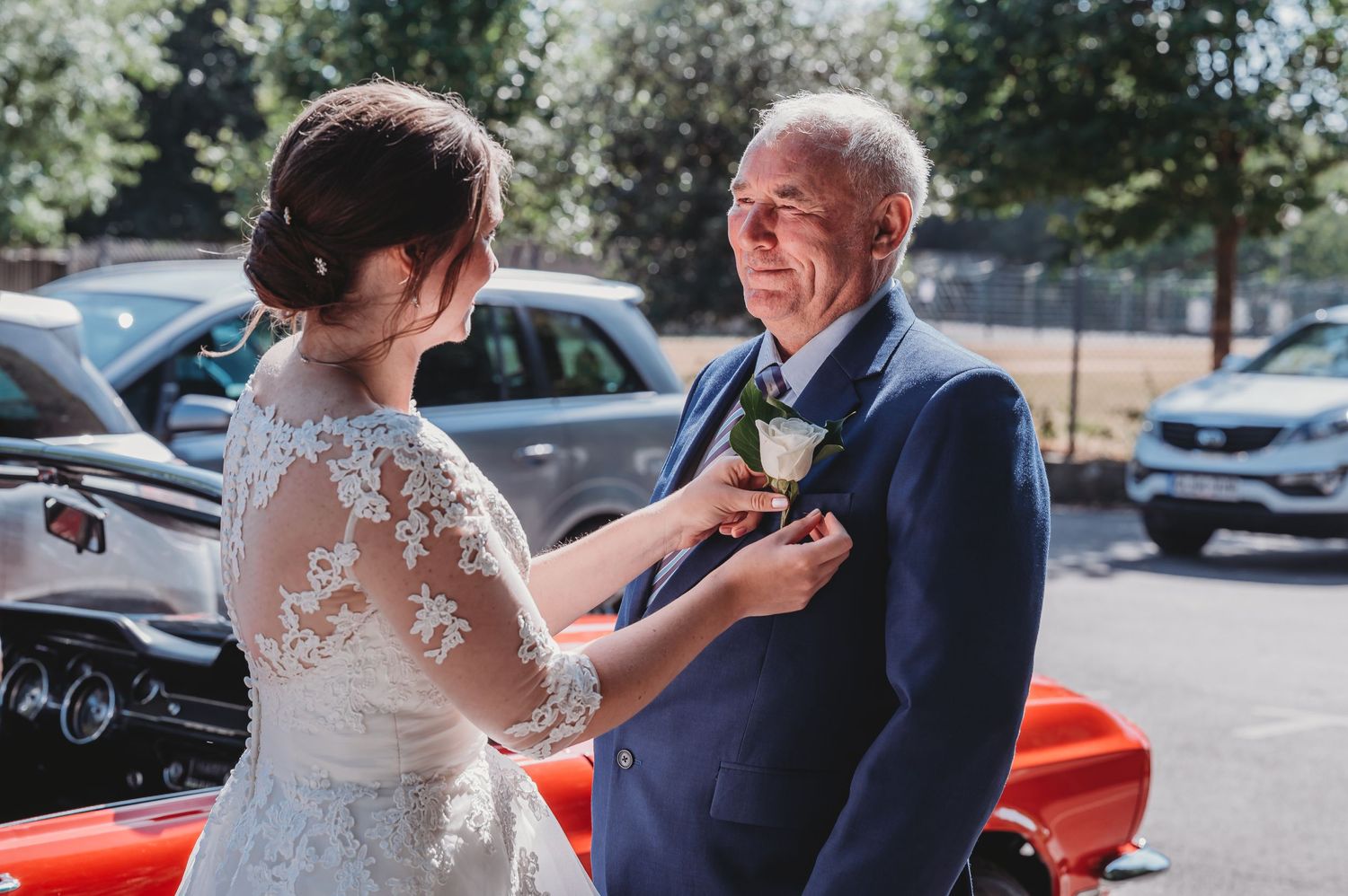 A bride adjusts a floral boutonniere on an older man wearing a navy suit in a sunny parking lot.