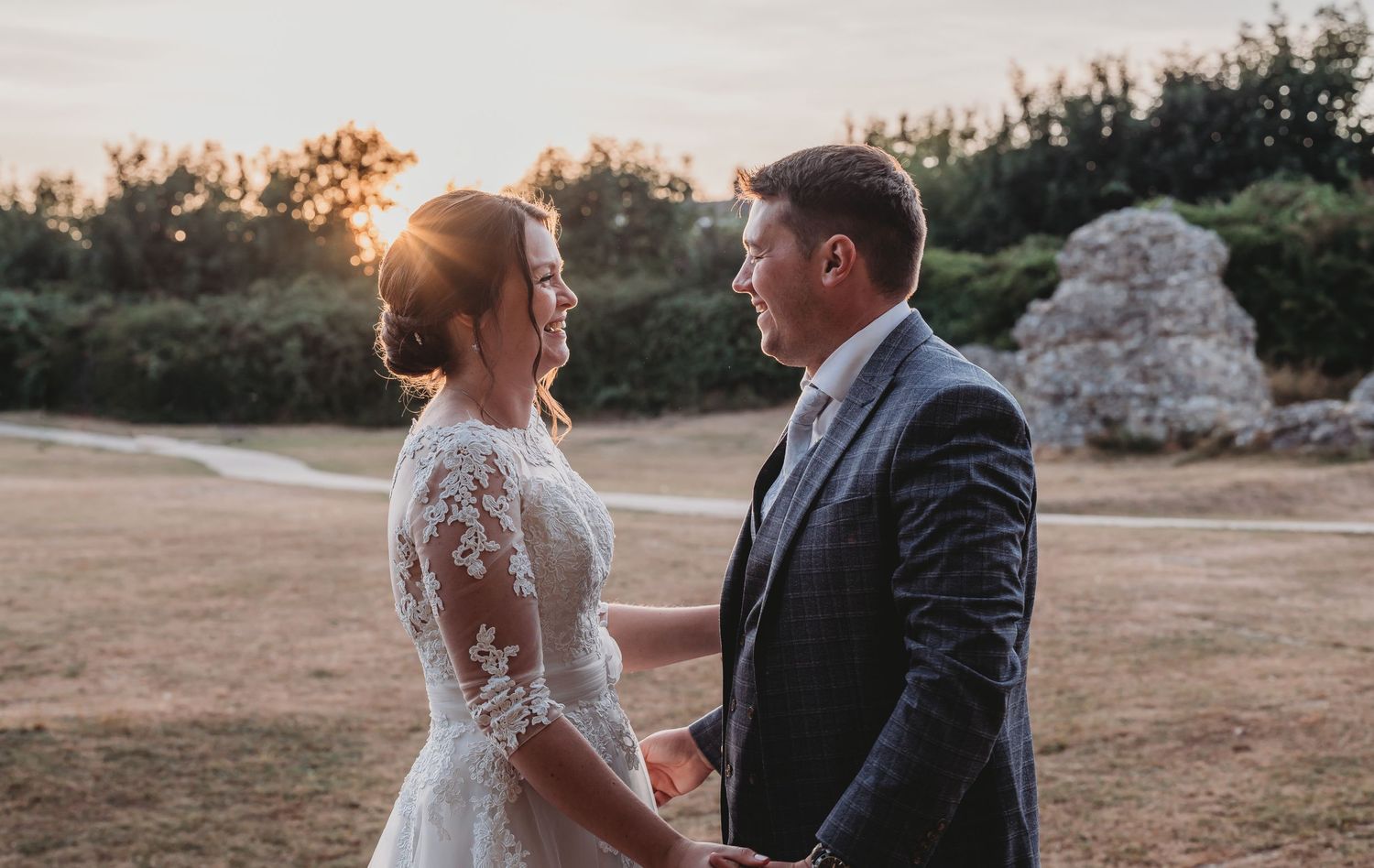 Couple holds hands at sunset during an outdoor wedding ceremony with lace dress and formal attire.
