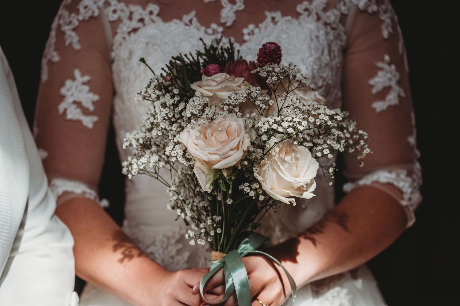 A delicate bridal bouquet of blush pink roses and baby's breath against a white lace dress background.