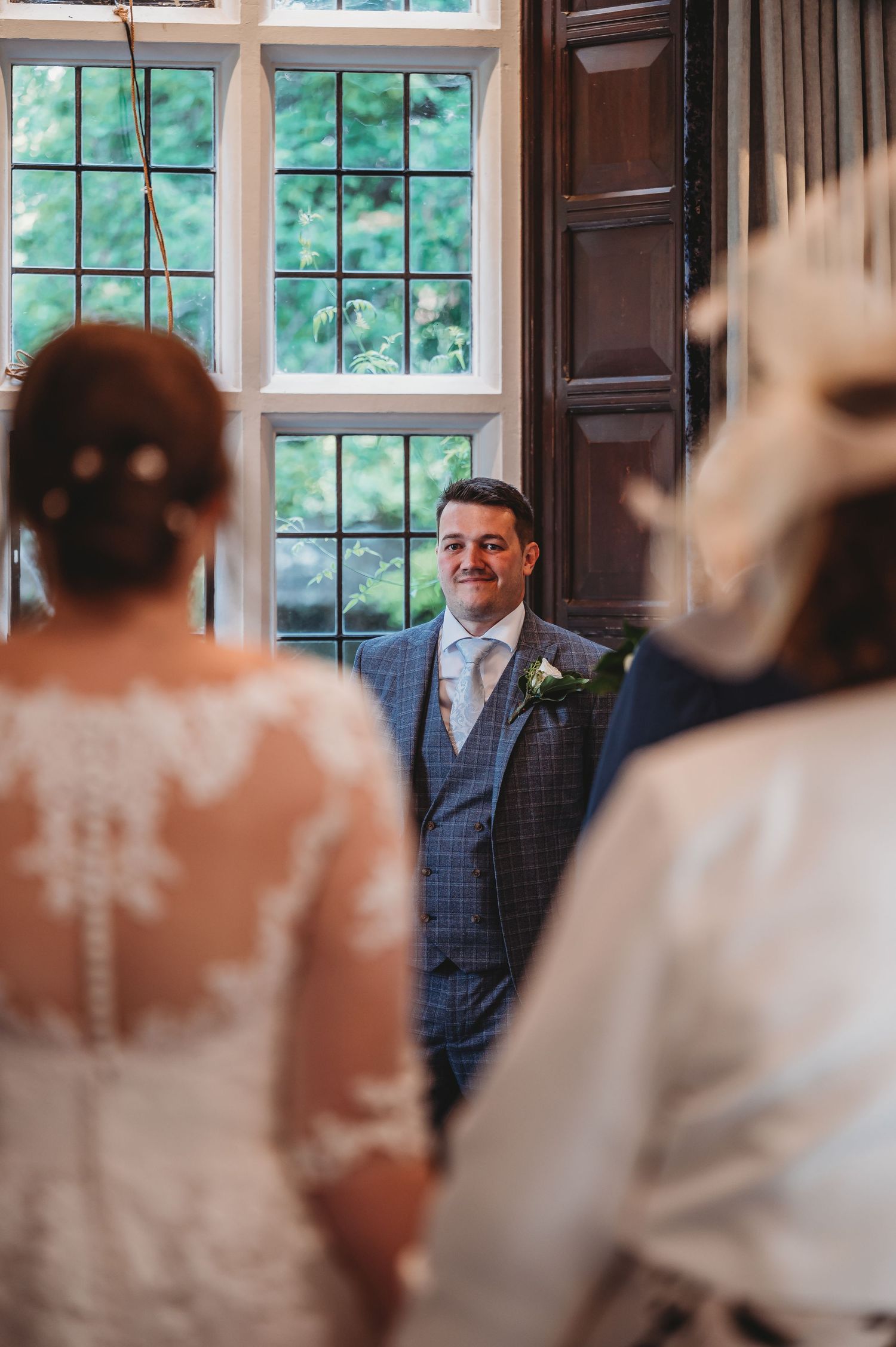 A groom in a blue checkered suit smiles during a wedding ceremony by a bright window.