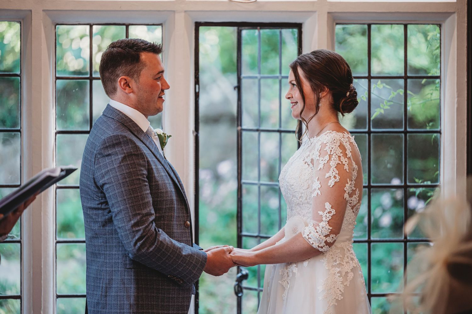 A couple holds hands during their wedding ceremony in front of sunlit windows in an indoor venue.