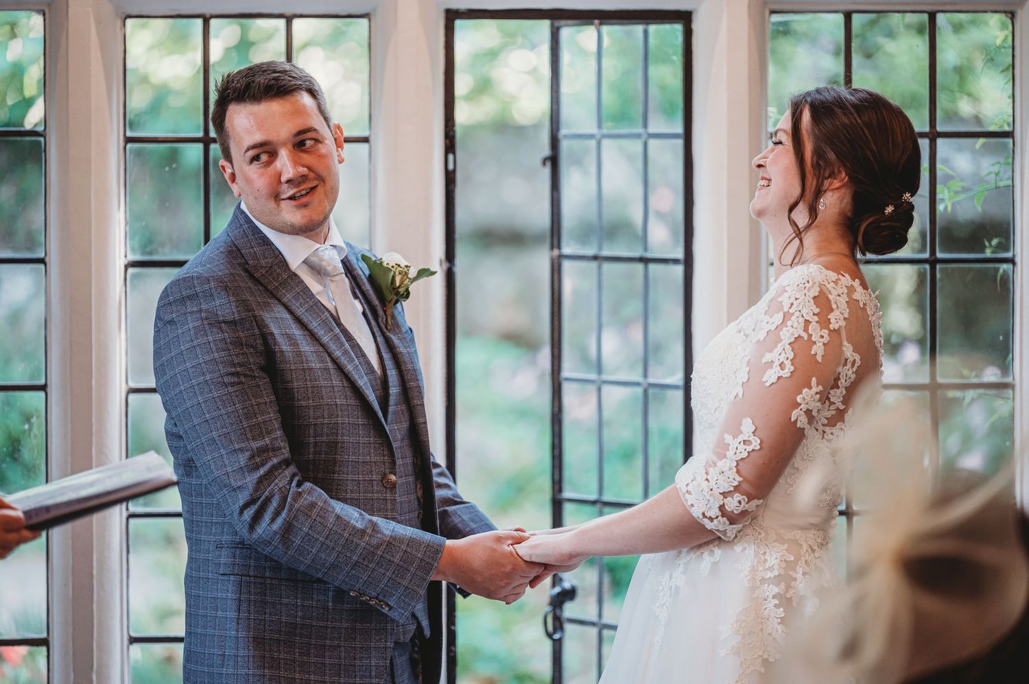 A couple stands together at their indoor wedding ceremony in front of glass windows.