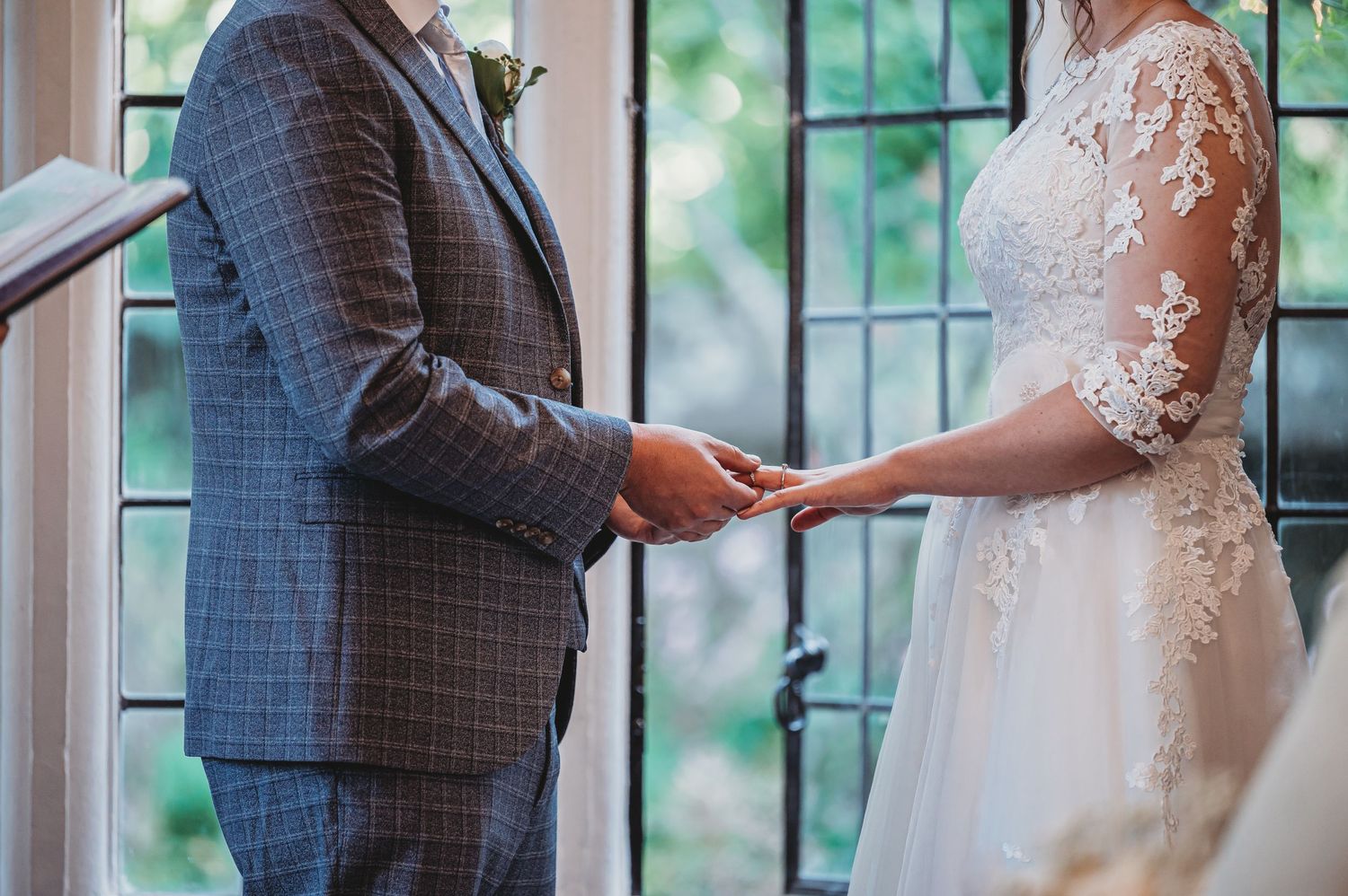 A couple exchanges wedding rings during an intimate ceremony beside a window with floral lace details on the dress.