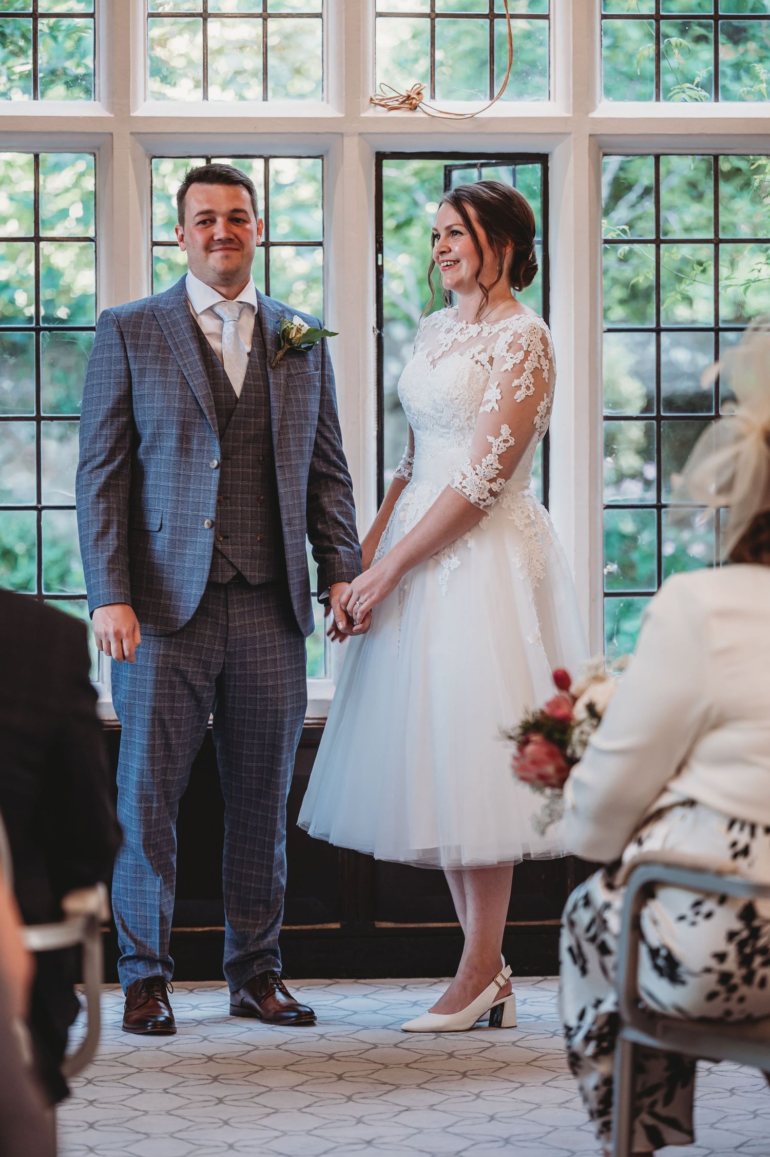 A wedding couple stands in front of a bright window with the bride wearing a tea-length dress and groom in a plaid suit.