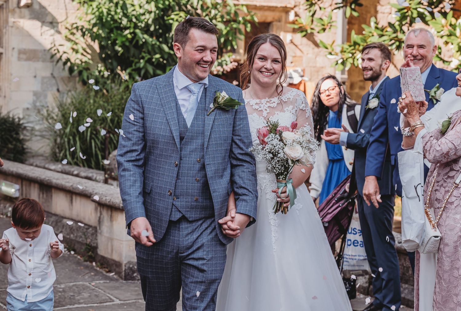 A newlywed couple walks down steps together while wedding guests look on and smile from behind.
