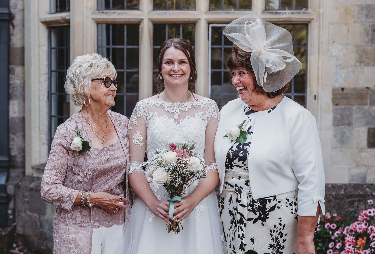 Elegant wedding gathering in formal attire with floral accents standing against stone building exterior.
