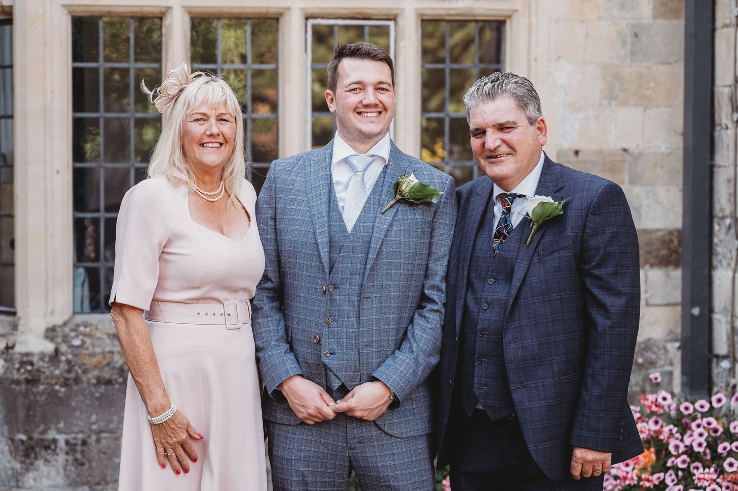 Three people pose for a formal family photo at a wedding outside a stone building with windows.