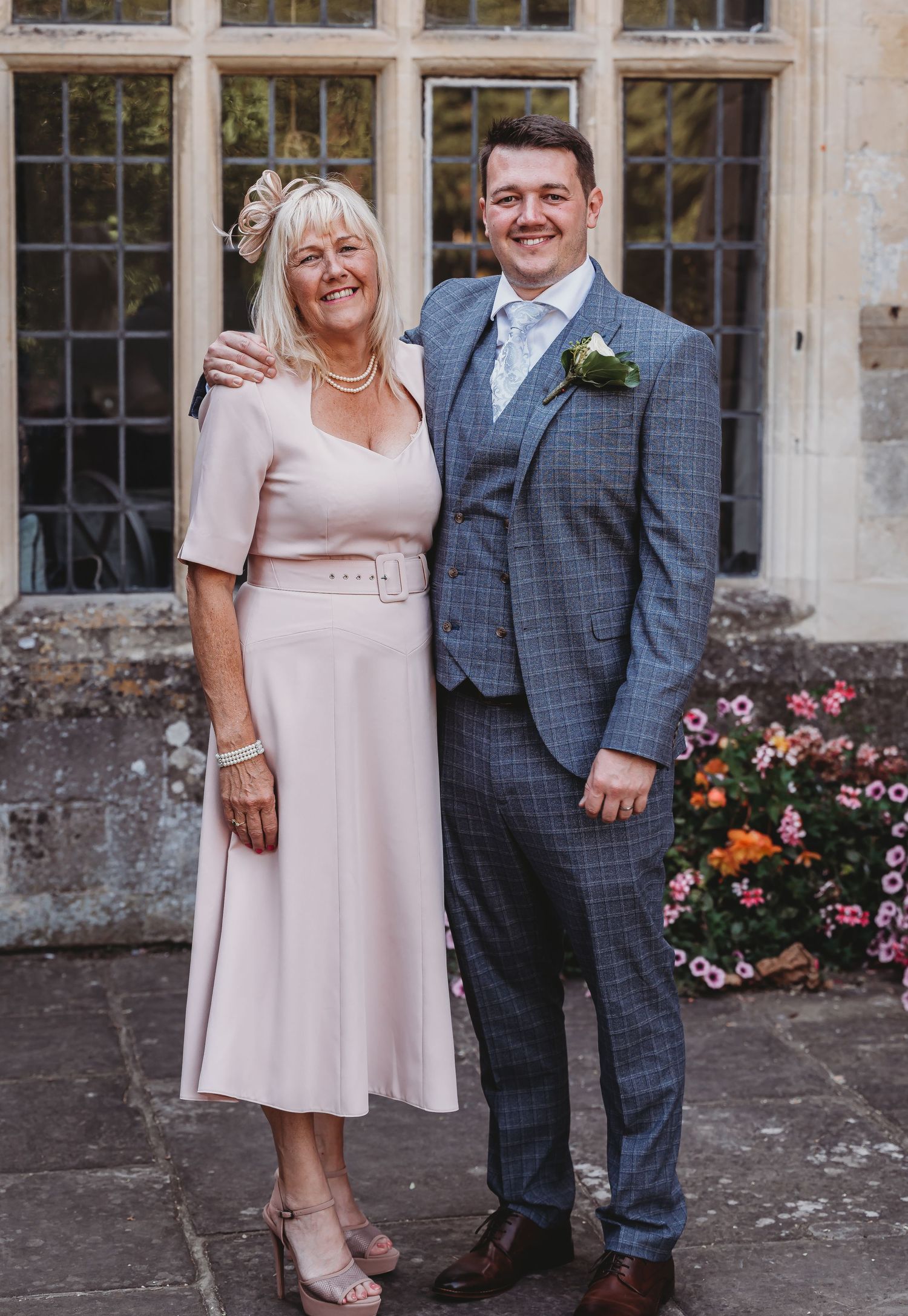 Well-dressed pair pose together at a formal event, with stone walls and windows in background, woman in pink dress.