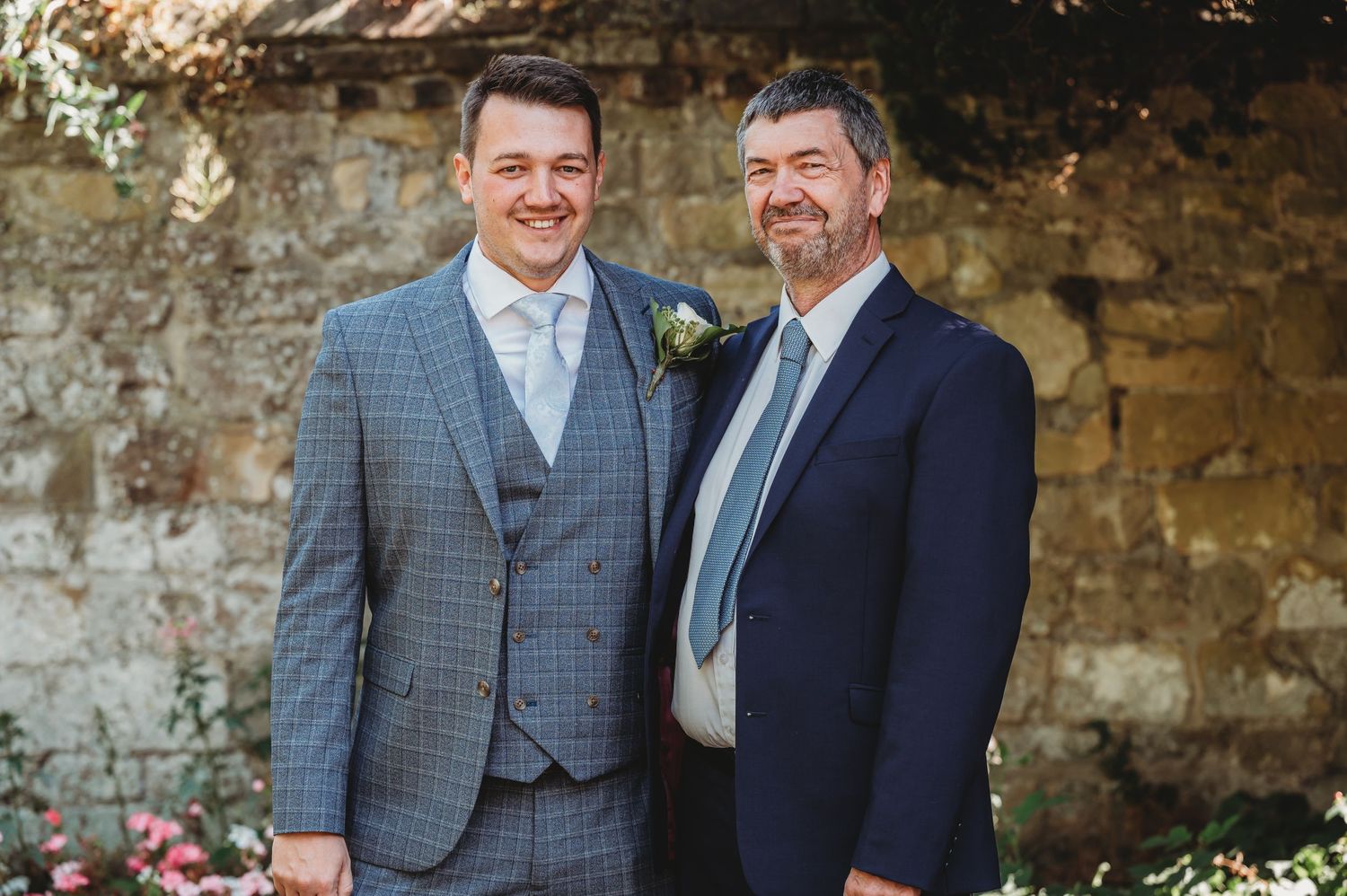 Two people in formal attire standing together at a rustic stone wall wearing suits and ties.