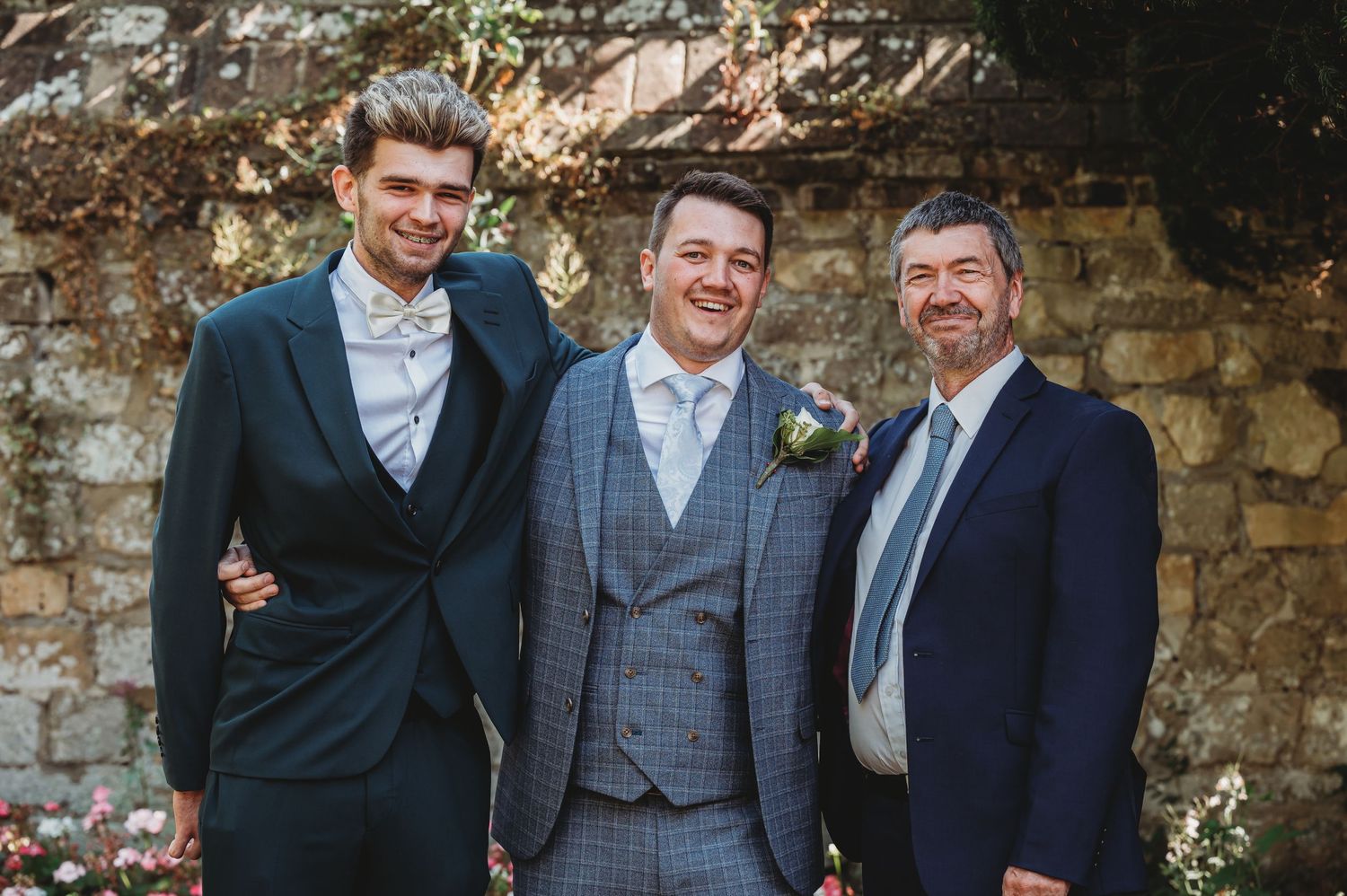 Three men in formal suits stand together smiling against a rustic stone wall backdrop at a wedding celebration.