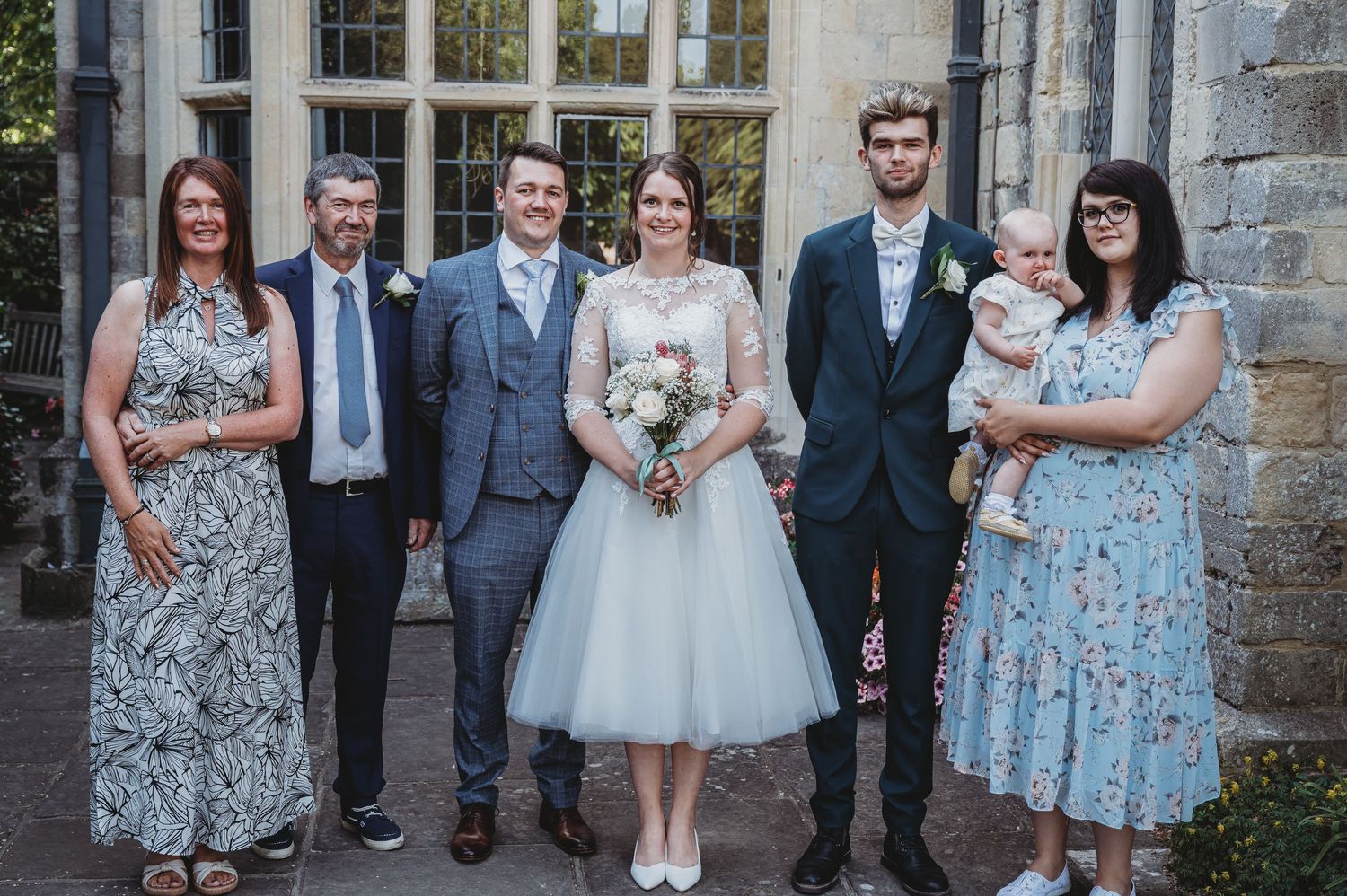 A small wedding party in formal attire stands together outside a stone building with windows.