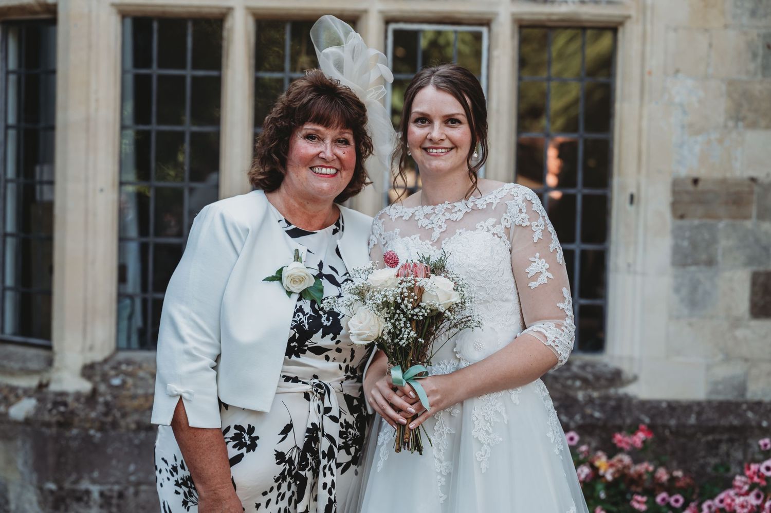 A bride wearing a white lace wedding dress holds a rustic bouquet while standing next to a guest in a floral dress.