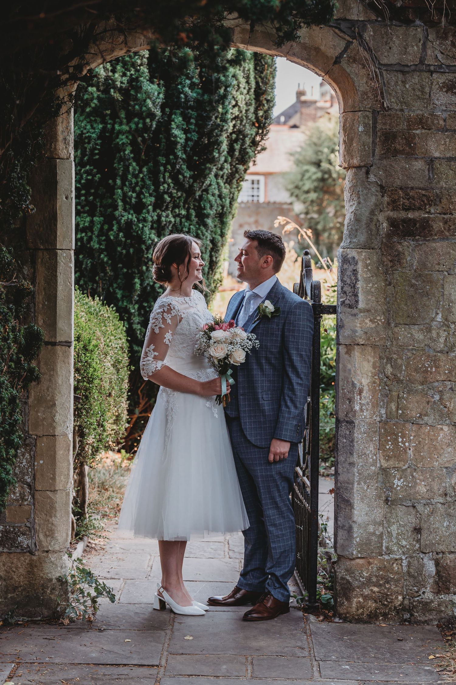 A wedding couple stands in a romantic stone archway with ivy, the bride wearing a tea-length dress and holding a bouquet.