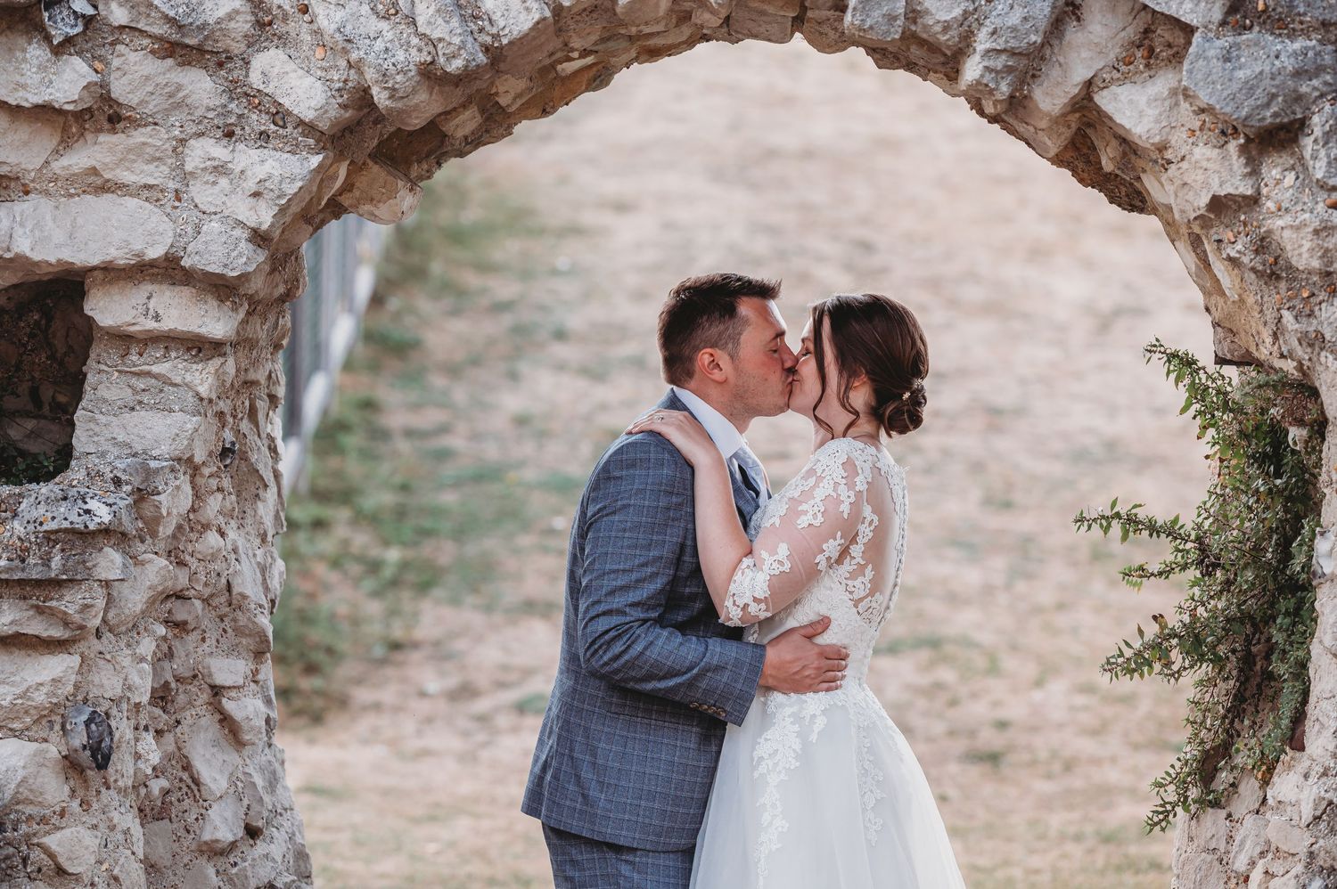 A romantic wedding couple embraces and kisses under a rustic stone archway in soft afternoon light.