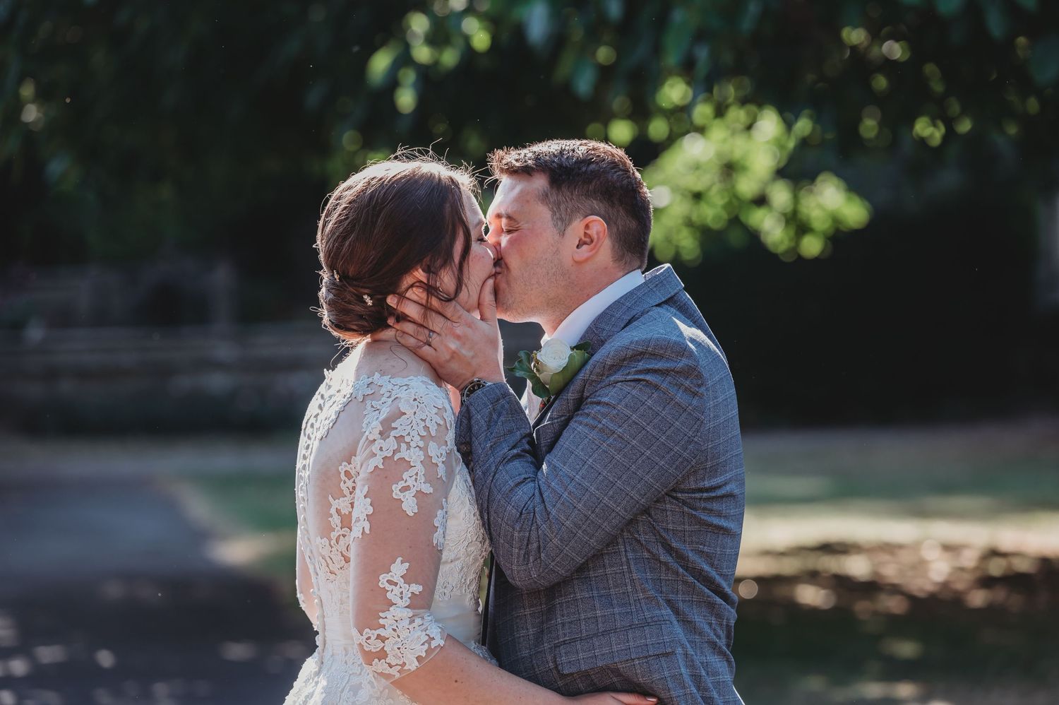 A couple shares a romantic kiss in an outdoor setting, with the bride wearing a lace dress and groom in a gray plaid suit.
