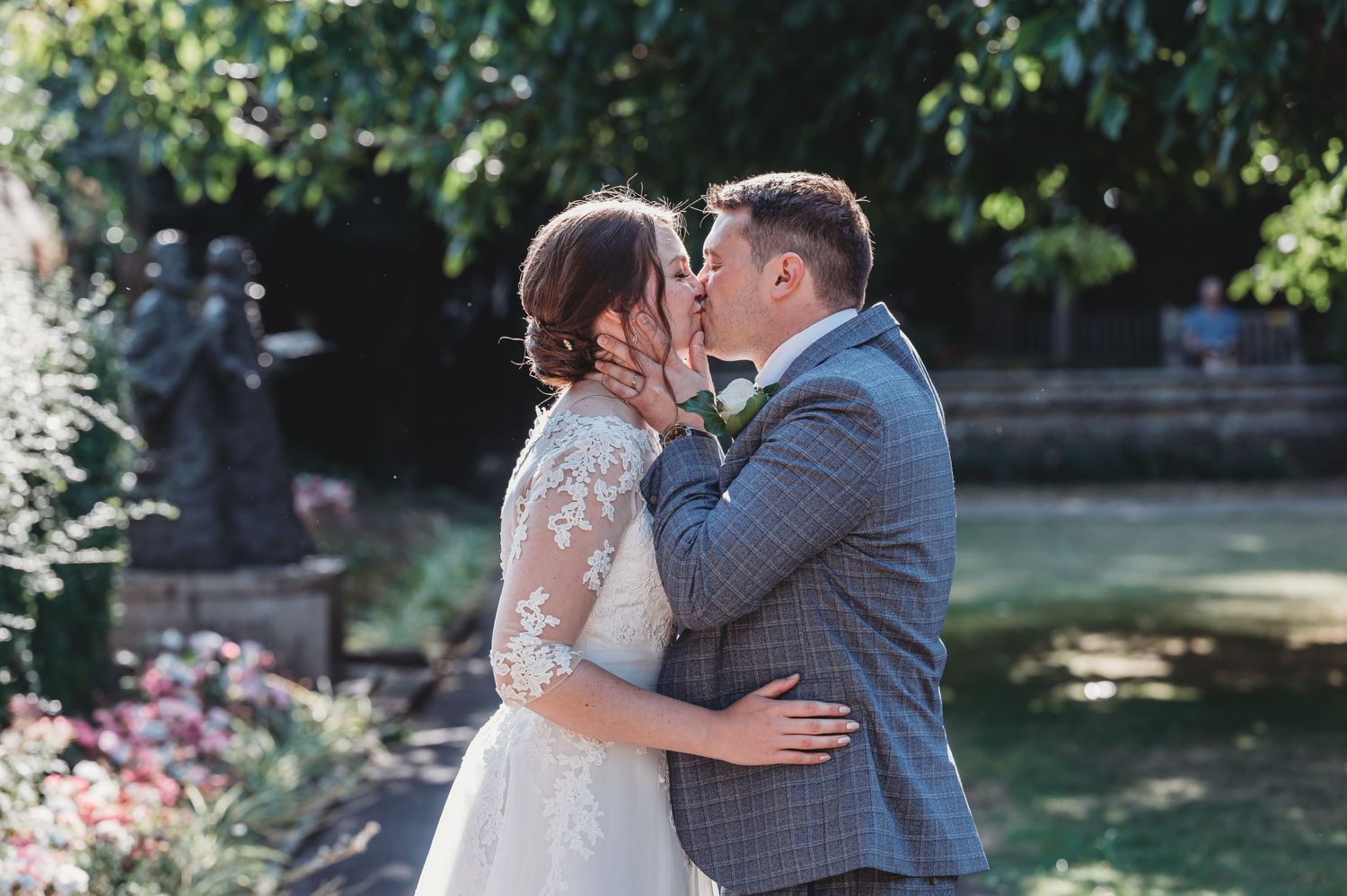 A romantic wedding couple shares a tender kiss in a garden setting with blooming flowers and sunlit greenery.