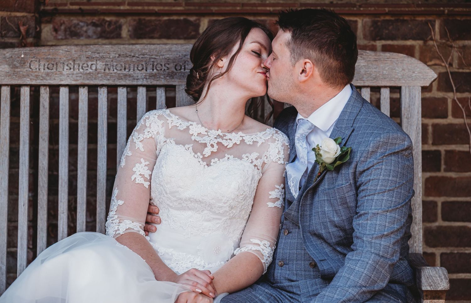 A tender wedding moment between a bride in lace sleeves and groom in gray suit sitting against brick wall.