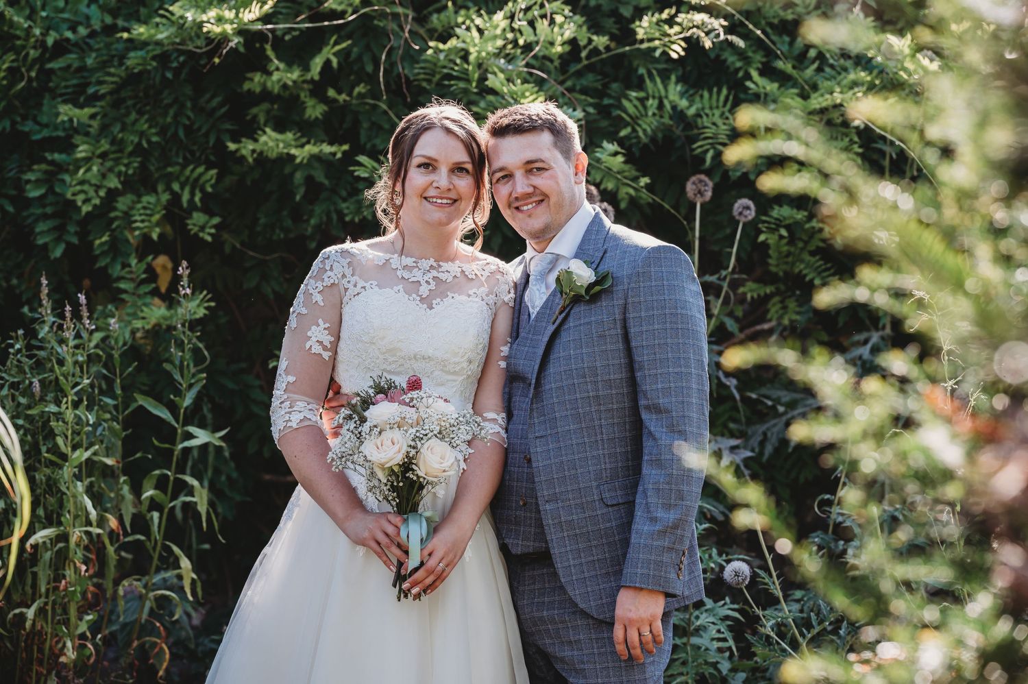 A couple poses together outdoors on their wedding day wearing formal attire with green foliage in the background.