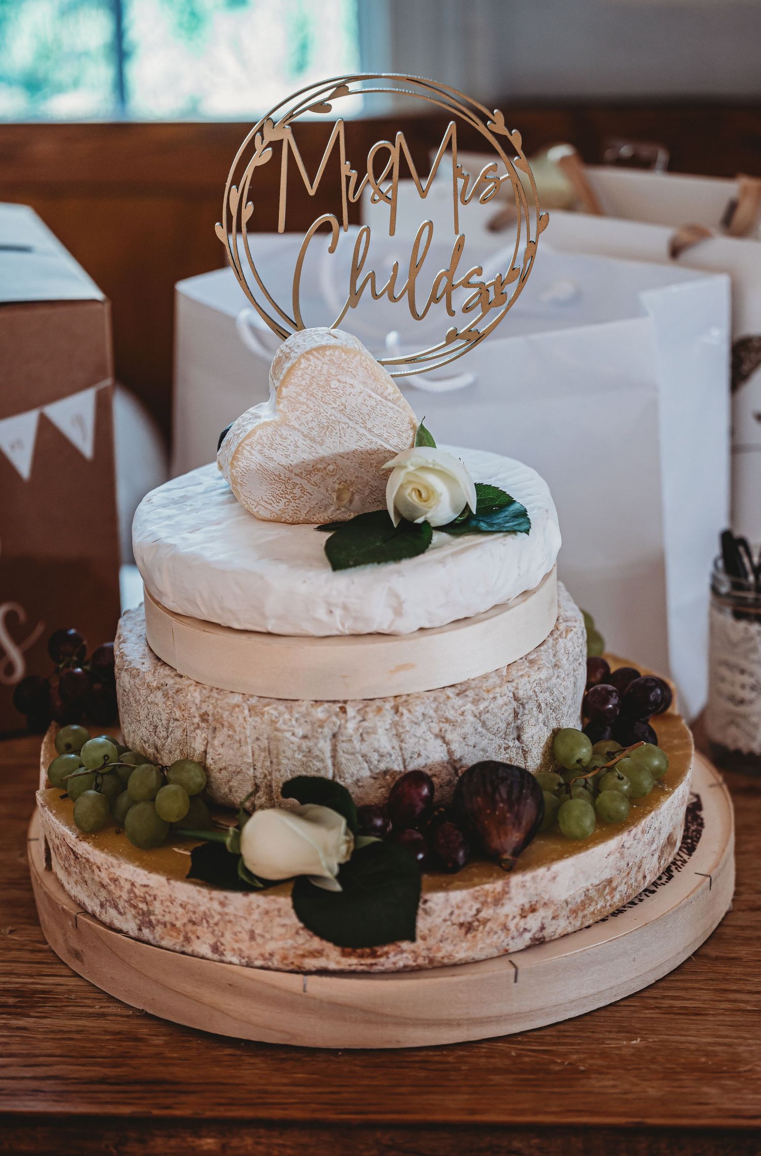 Wedding cheese wheel cake decorated with grapes, figs, and gold 'Mr & Mrs' topper on wooden table.