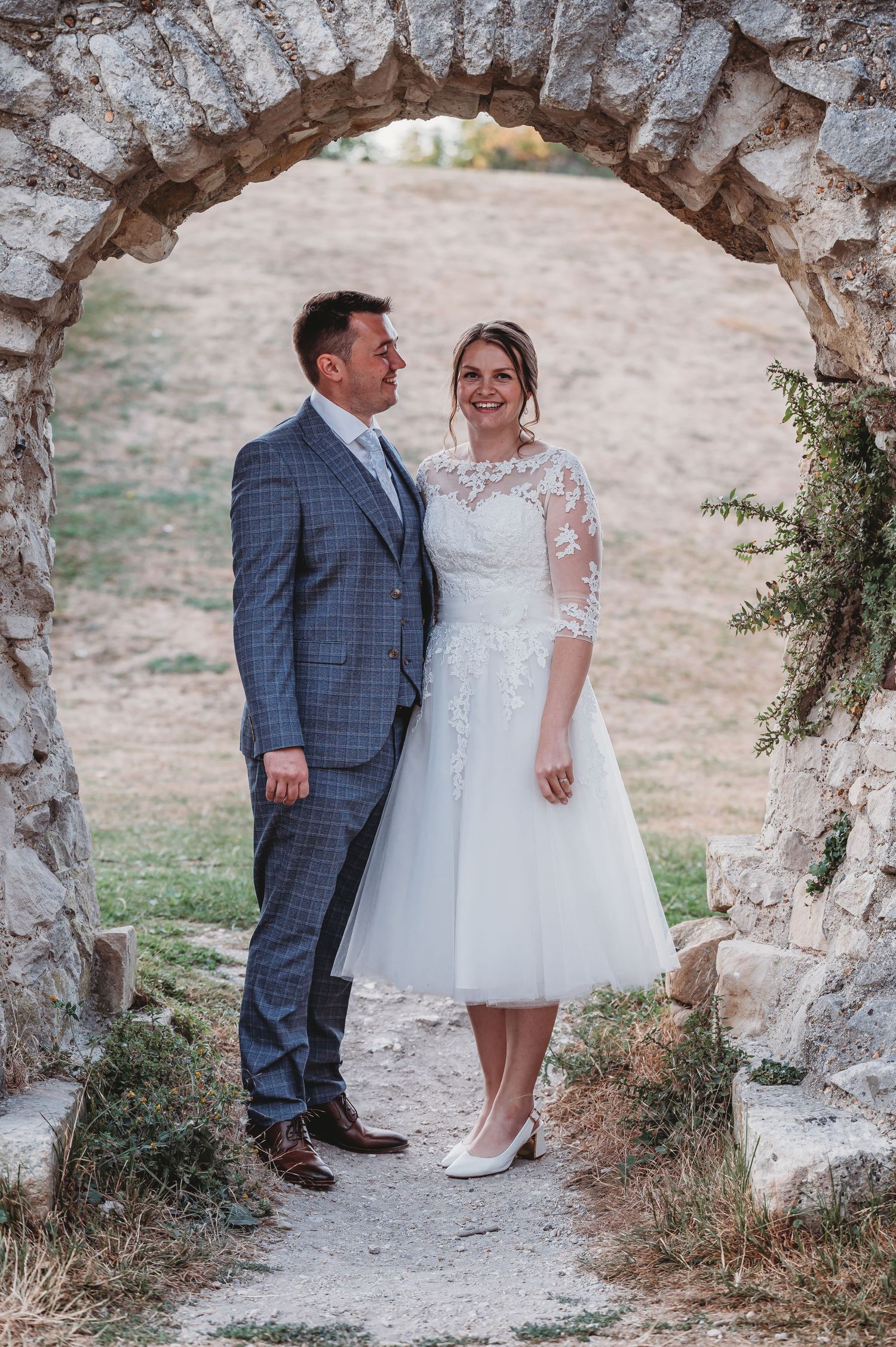 A bride and groom pose together under a rustic stone archway during their romantic countryside wedding.