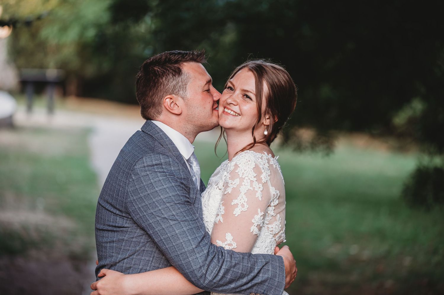 A romantic wedding couple embraces outdoors with the bride wearing a lace dress and groom in grey suit.