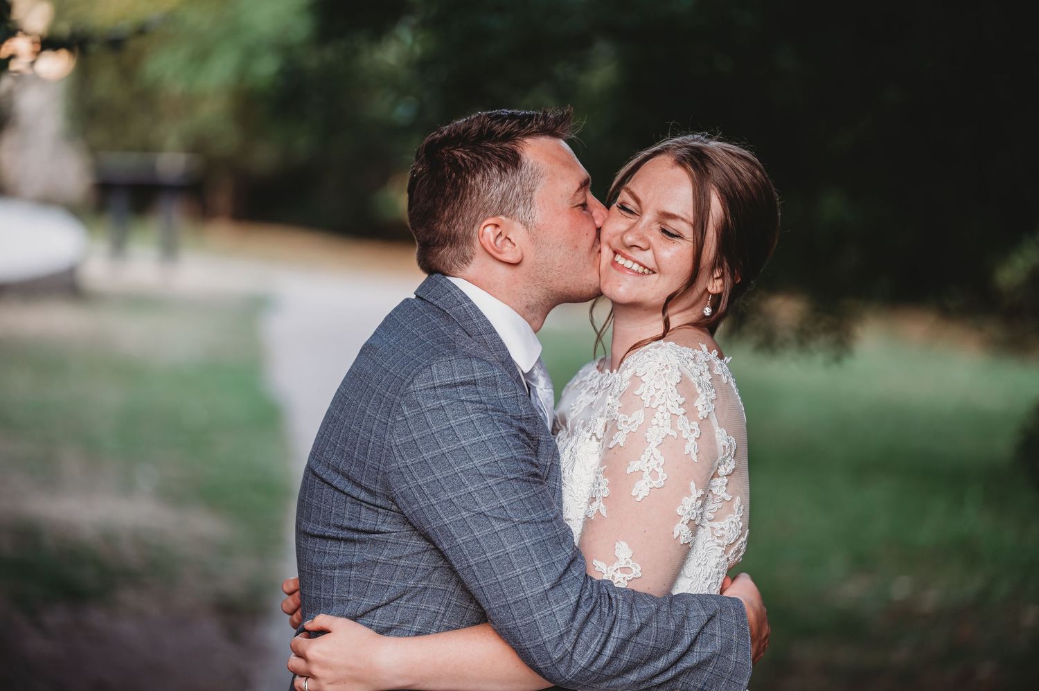 A romantic embrace between newlyweds outdoors, with the bride wearing a lace dress and groom in a gray check suit.