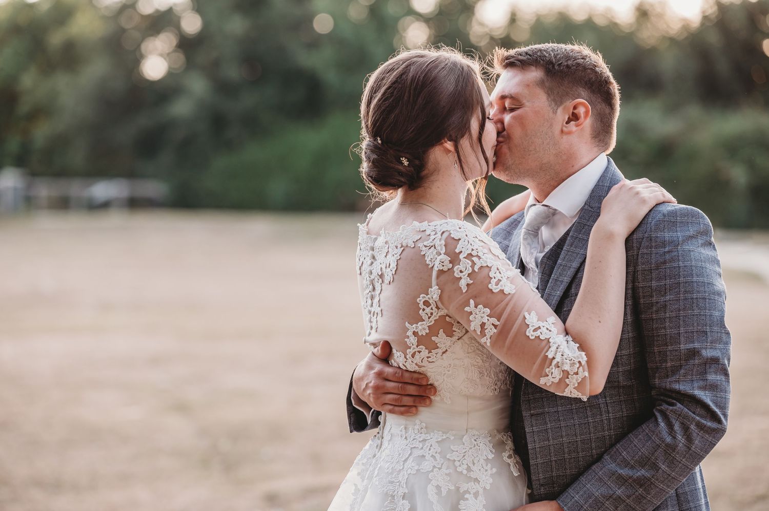 A romantic wedding couple shares a tender kiss at sunset in an outdoor setting, her lace dress glowing in golden light.