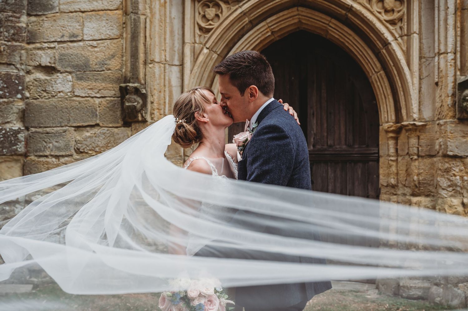A romantic wedding couple kisses in front of an old stone church archway as the bride's long veil flows in the wind.