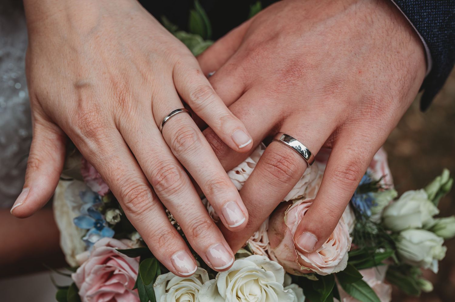 Close up of bride and groom's hands showing wedding rings against pink and white roses.