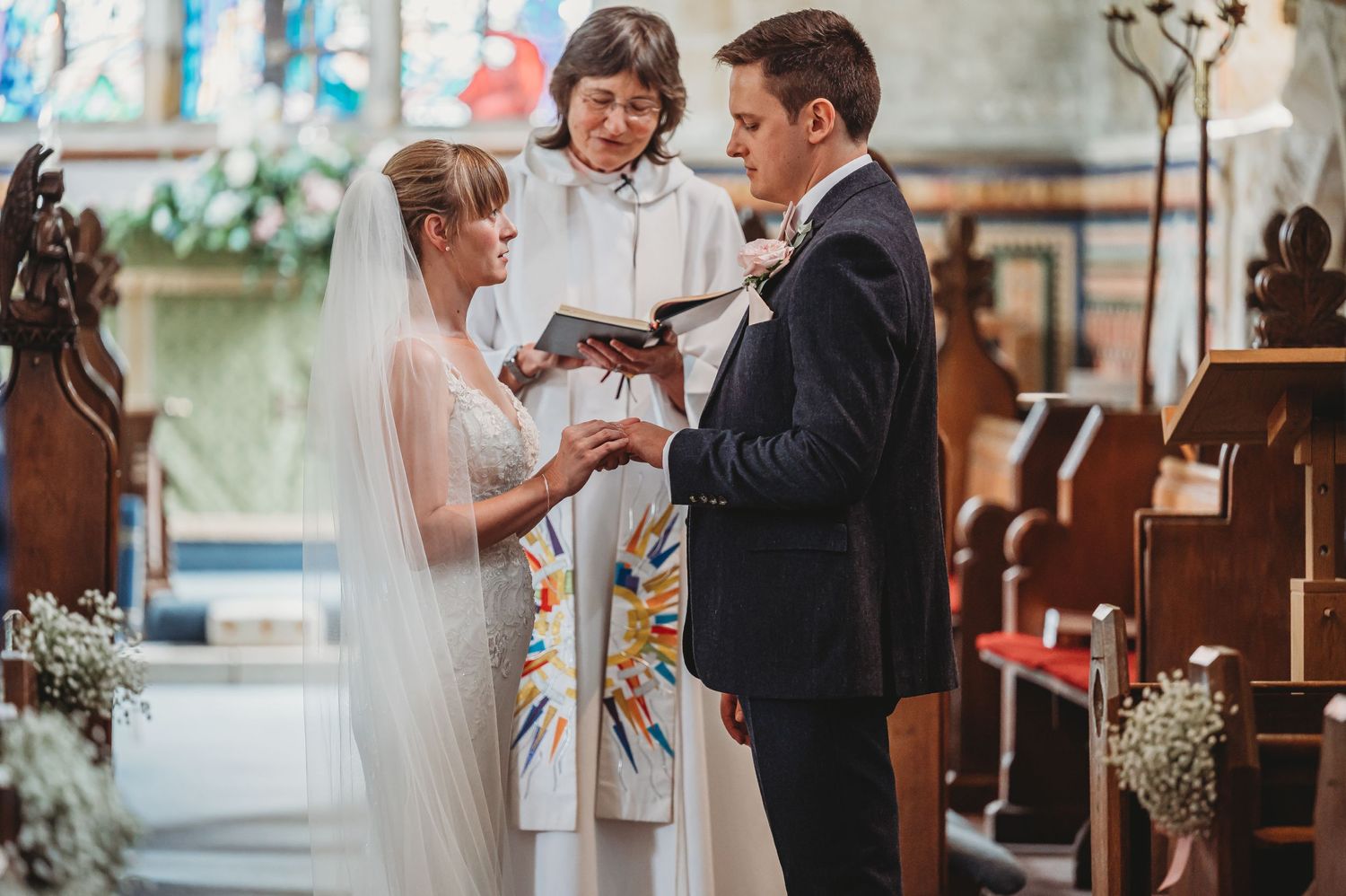 Two people exchange rings during an intimate wedding ceremony in a church with wooden pews and floral decorations.