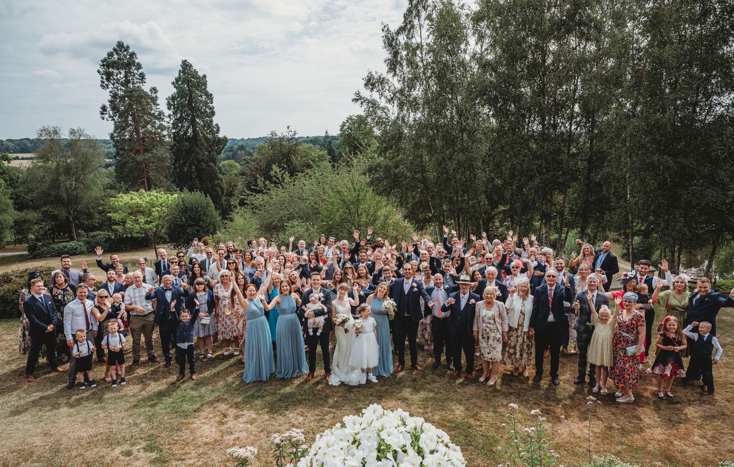 Large group of wedding guests gathered outdoors on a hillside with trees and floral arrangements for a scenic ceremony.