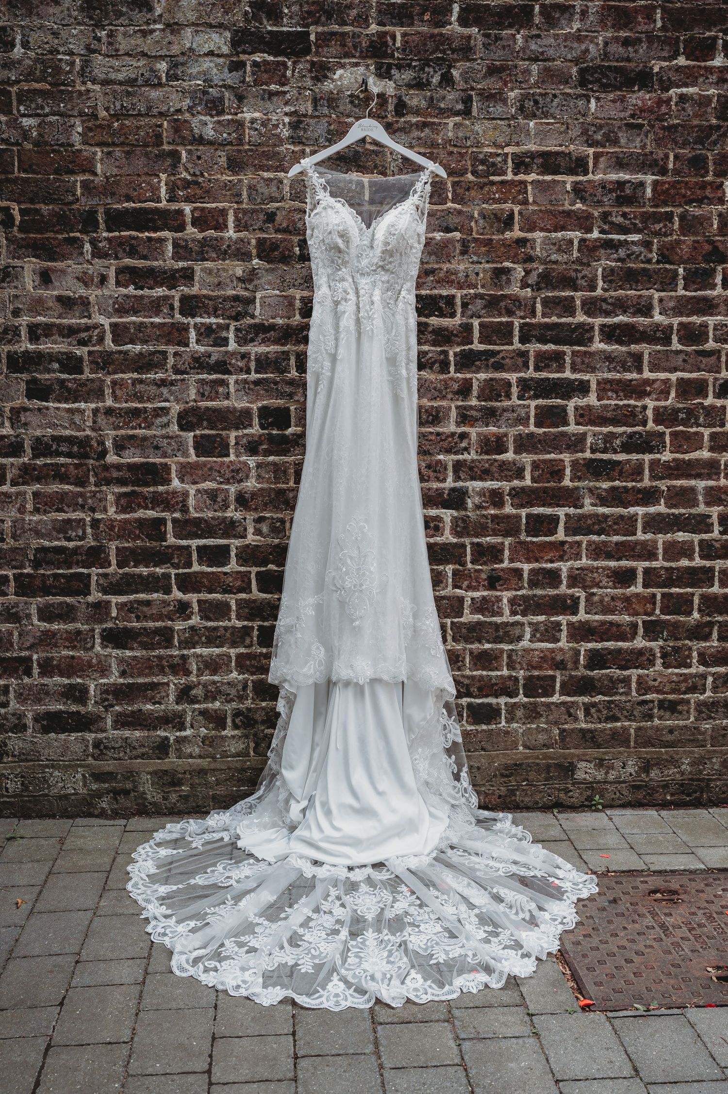 Elegant white lace wedding dress with sweetheart neckline and long train hanging against rustic brick wall.