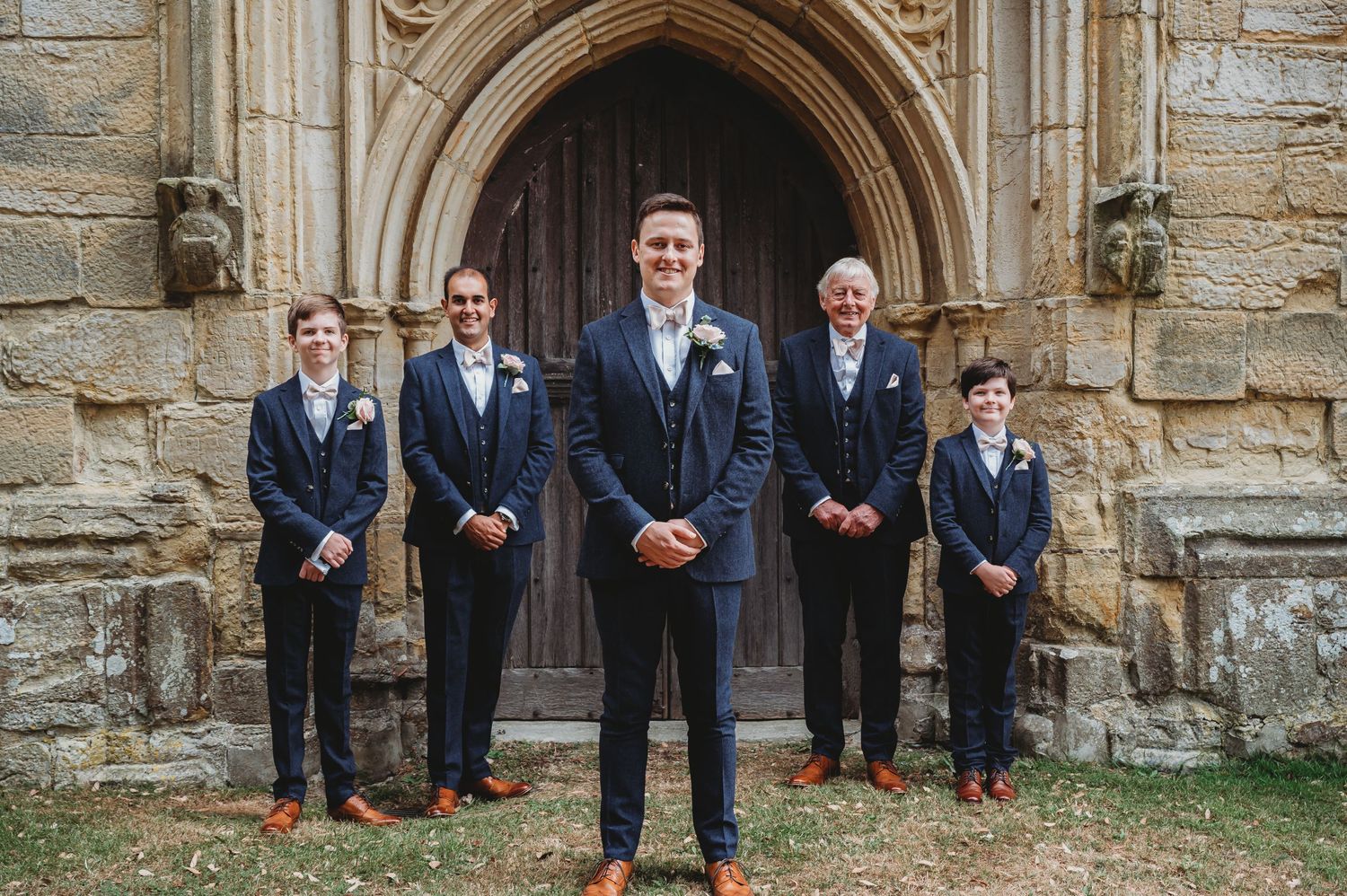 Five people in navy suits stand in formation in front of a gothic stone church doorway.