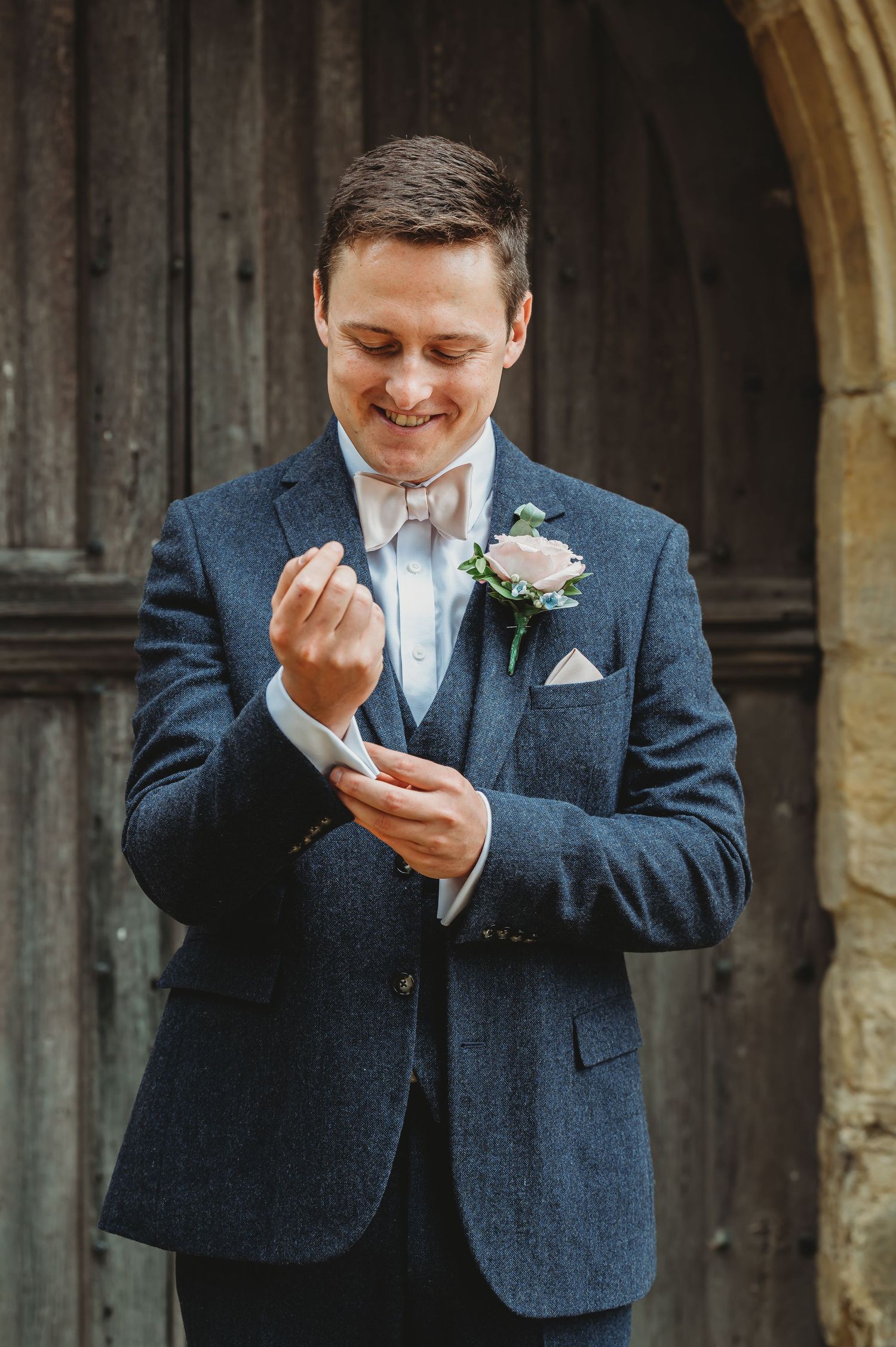 A groom in a navy tweed suit adjusts his cuff while smiling, wearing a pink bow tie and white boutonniere.