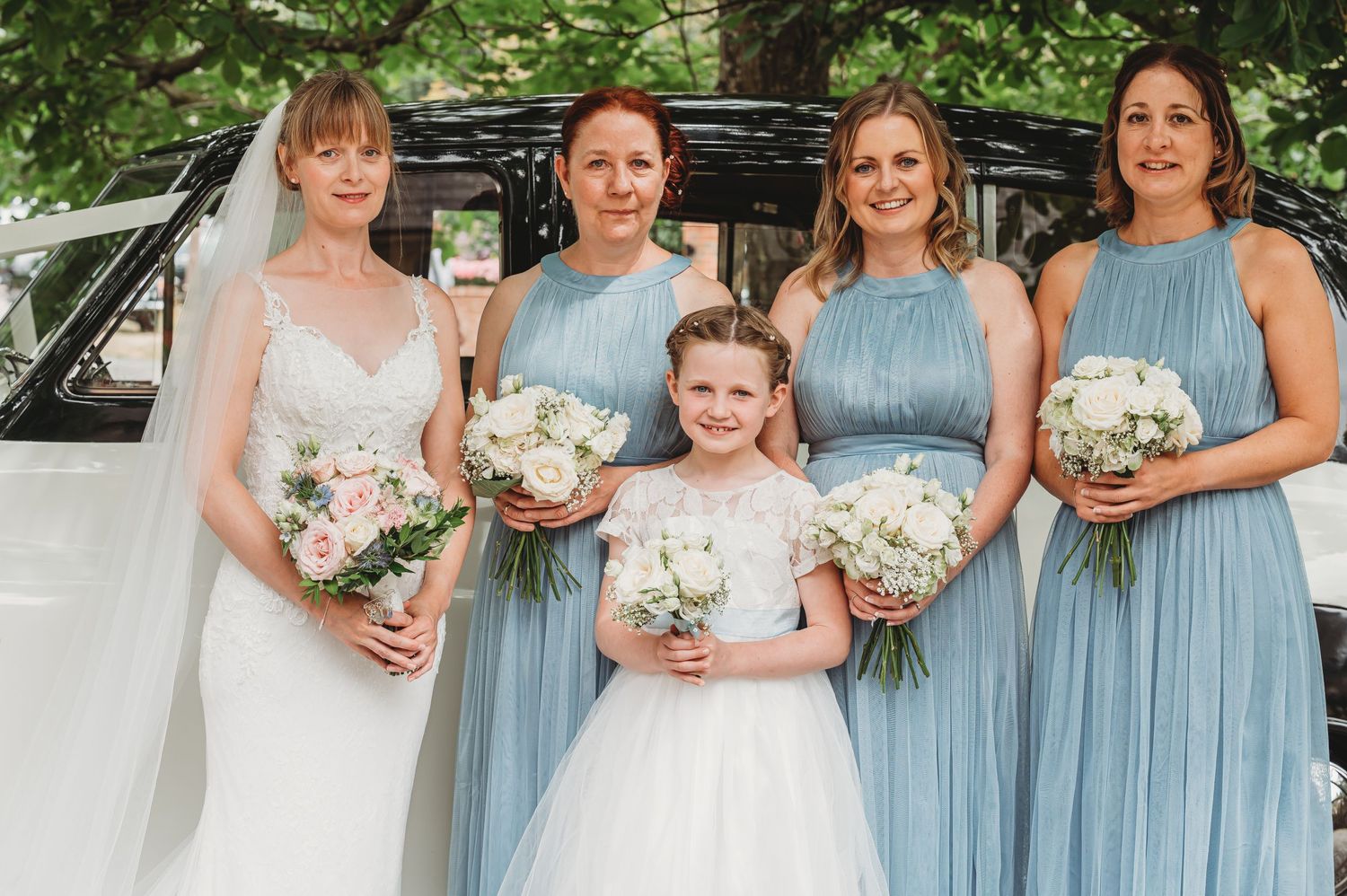 Wedding party in matching dusty blue bridesmaid dresses and white bouquets pose together outdoors by a car.