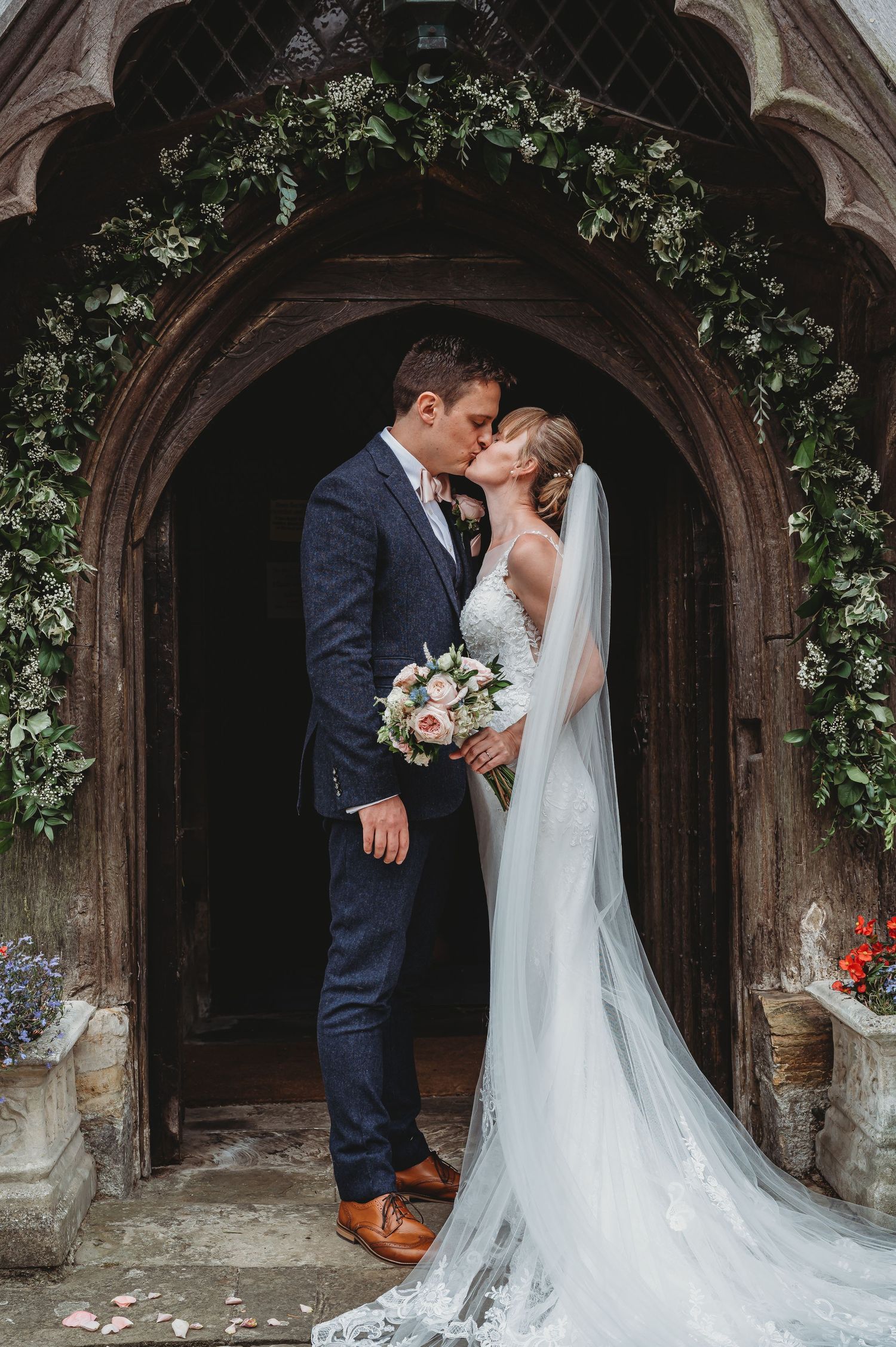 Newlywed couple shares romantic kiss in church doorway decorated with green floral arch and flowing white veil.