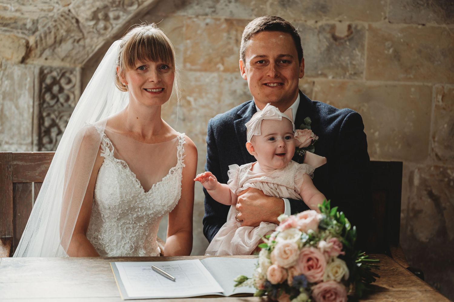 Newlyweds sign marriage documents at altar with baby while bouquet of pink roses rests on table.