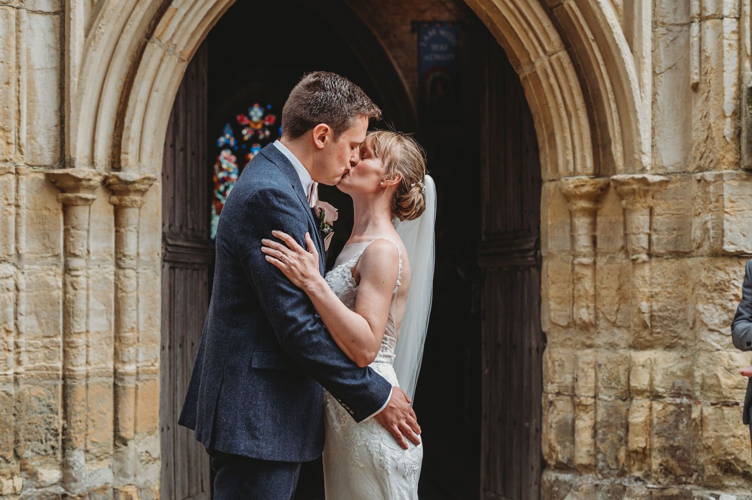 A couple shares a romantic kiss under a stone archway at a historic church building.