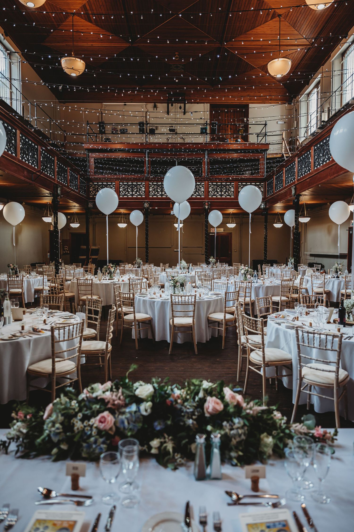 Elegant wedding reception venue with white round tables, string lights, floating white balloons and floral centerpieces.