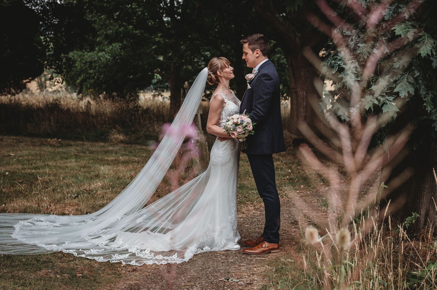 A romantic wedding photo captured outdoors with a long flowing white veil and natural landscape setting.
