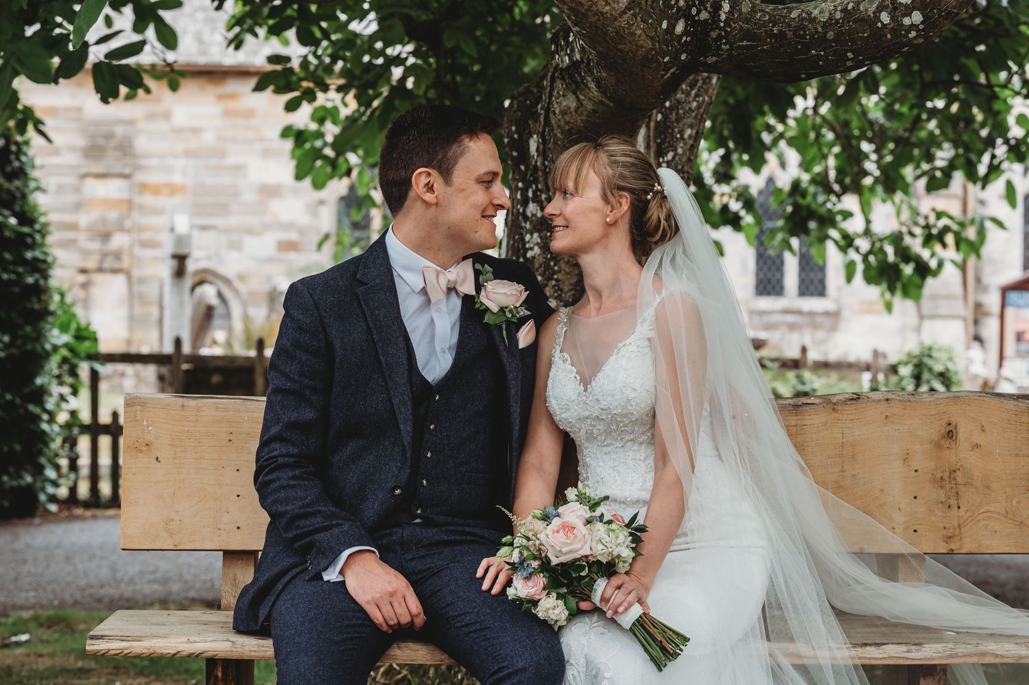 A bride and groom share an intimate moment while sitting on a bench under greenery at a rustic outdoor venue.