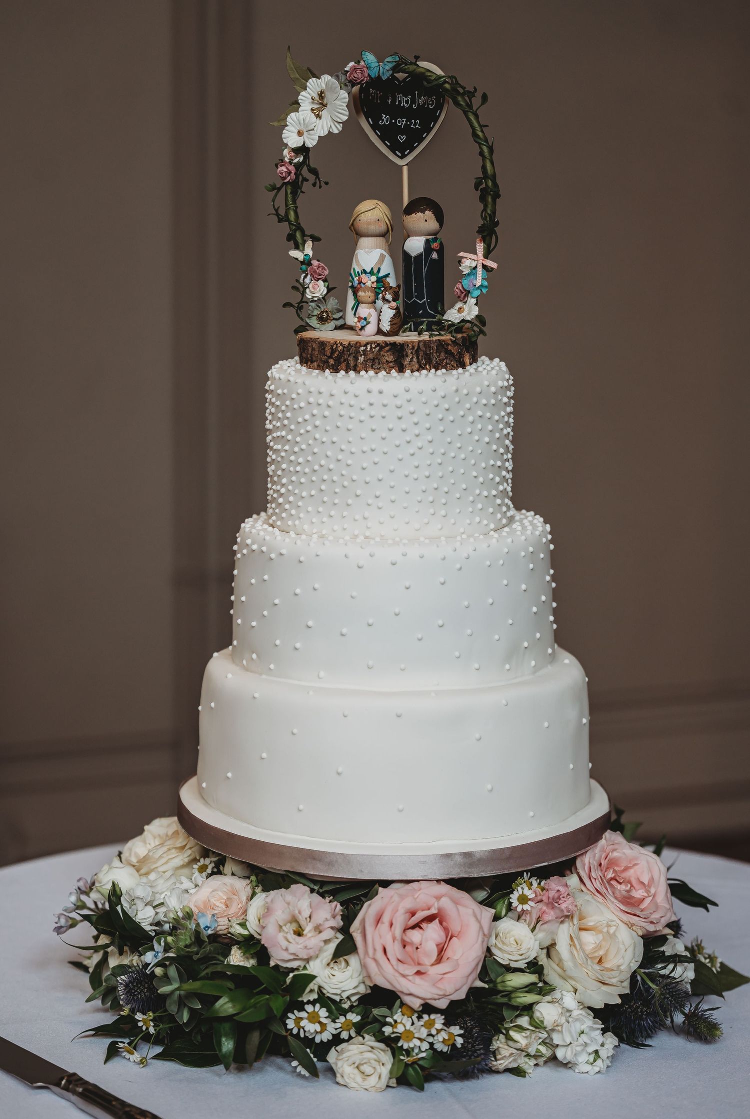 Elegant three-tiered white wedding cake with pearl details and floral topper surrounded by pink and cream roses.