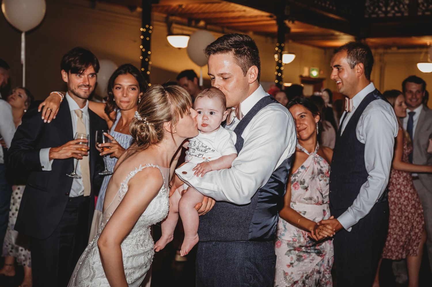 Couple dancing with a small child at an evening wedding reception while guests watch and celebrate in the background.