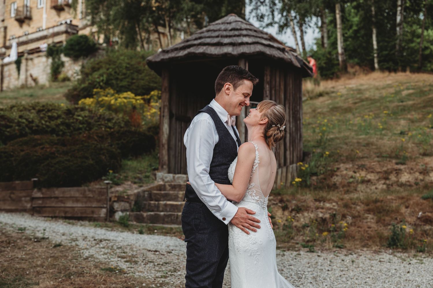 A romantic wedding couple embraces in front of a rustic wooden gazebo on a gravel path during an outdoor celebration.