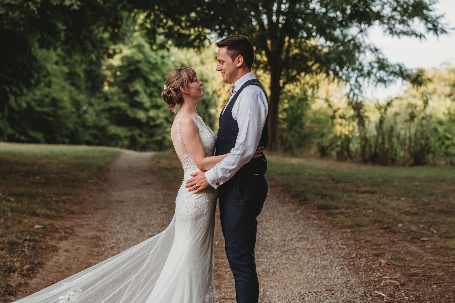 A bride and groom share an intimate moment on a rural dirt path surrounded by green trees and autumn foliage.