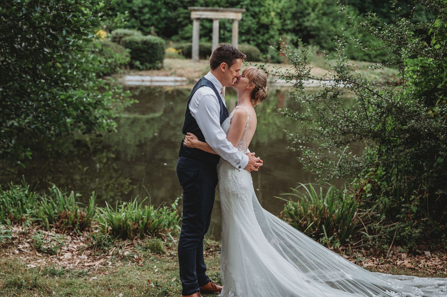 Couple in wedding attire embrace by a scenic pond with lush greenery and a stone structure in the background.