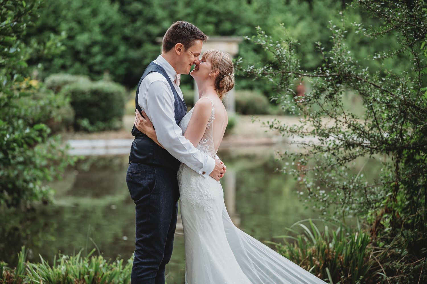 Two people share a romantic embrace near a lake in a lush garden setting, the bride wearing a flowing white gown.