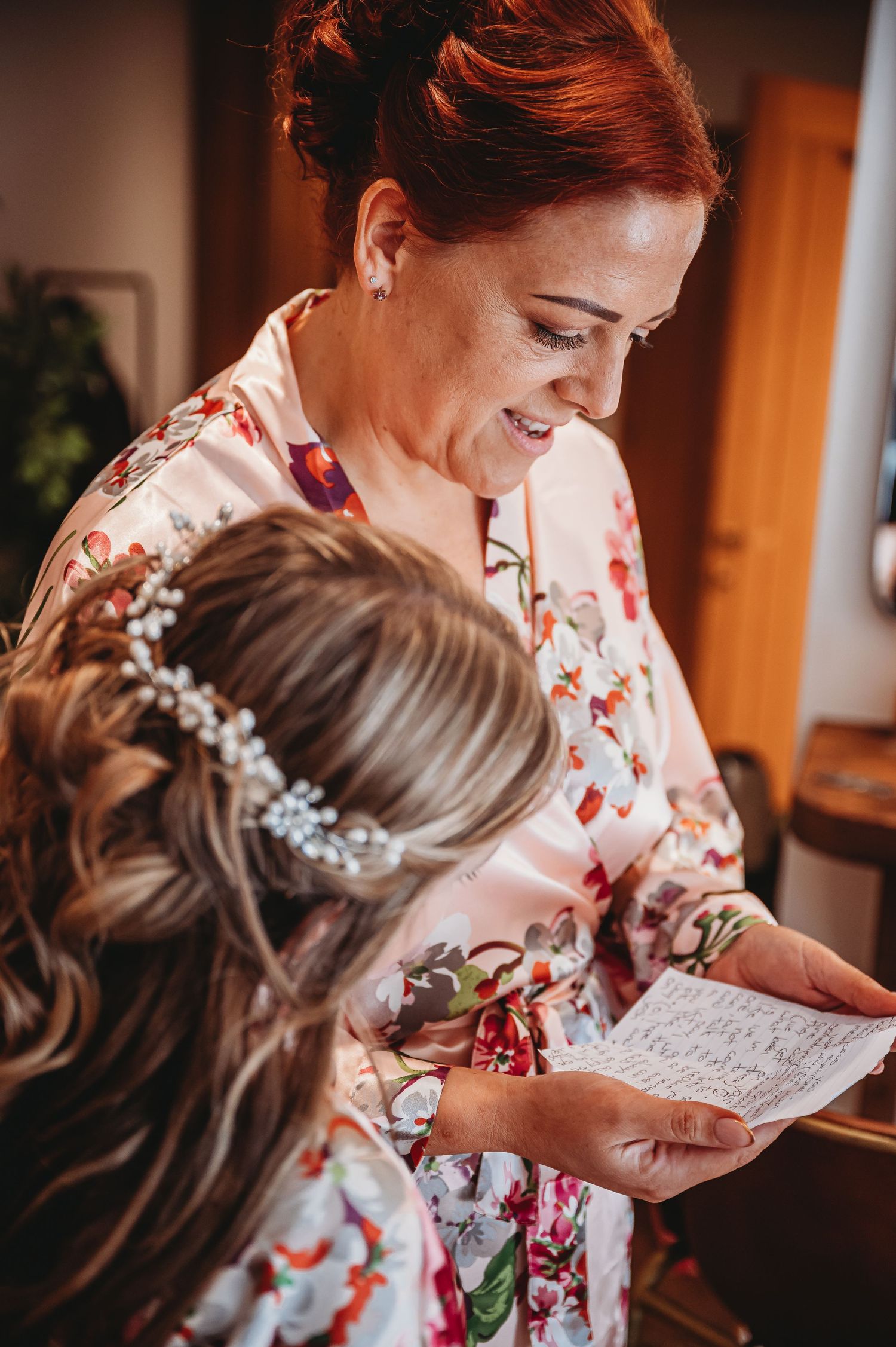 A woman in a floral robe smiles while reading a handwritten note, as someone with an elegant hairpiece looks on.