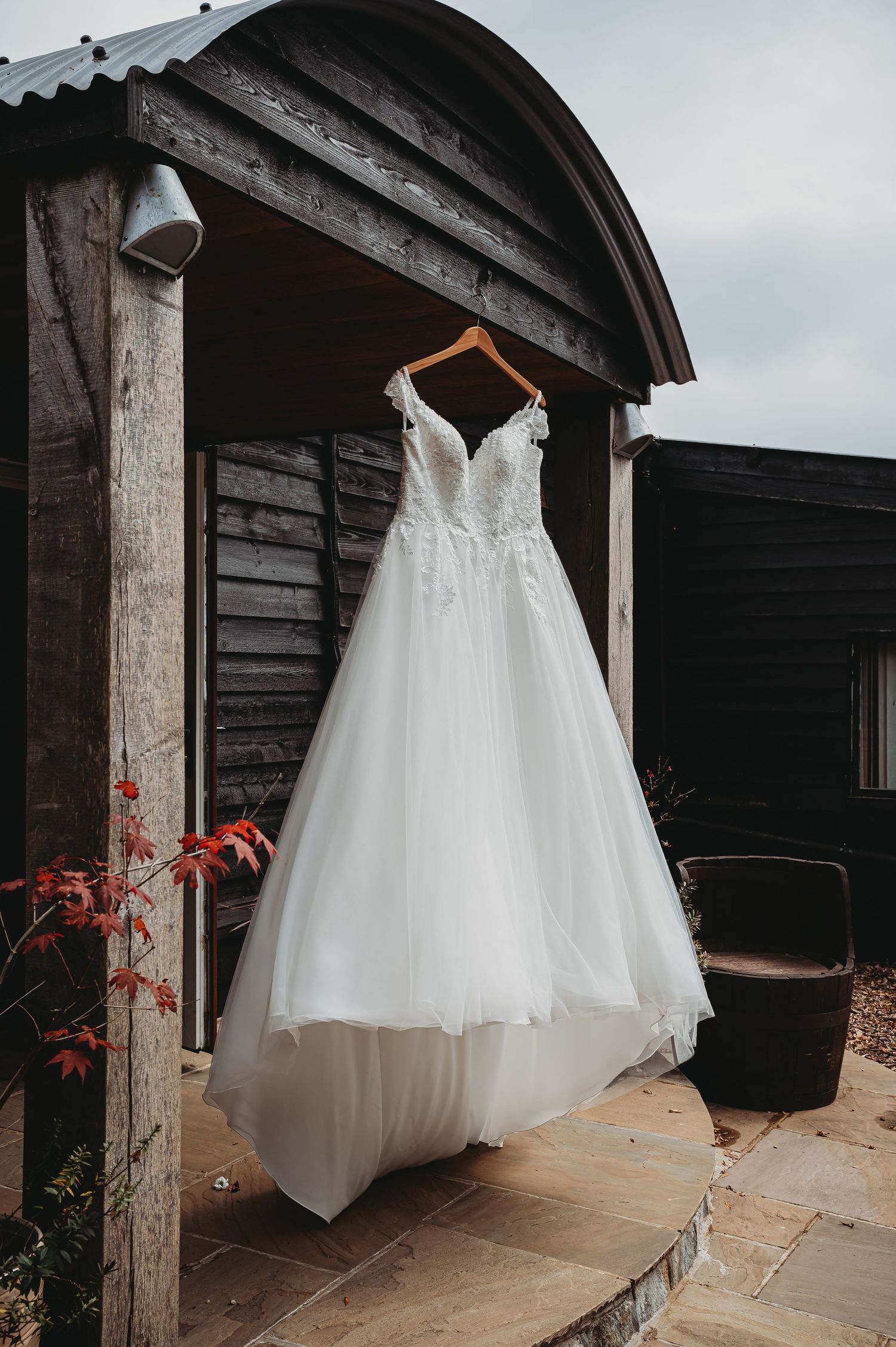 A white lace wedding dress hangs on a rustic wooden porch with red autumn leaves in the background.